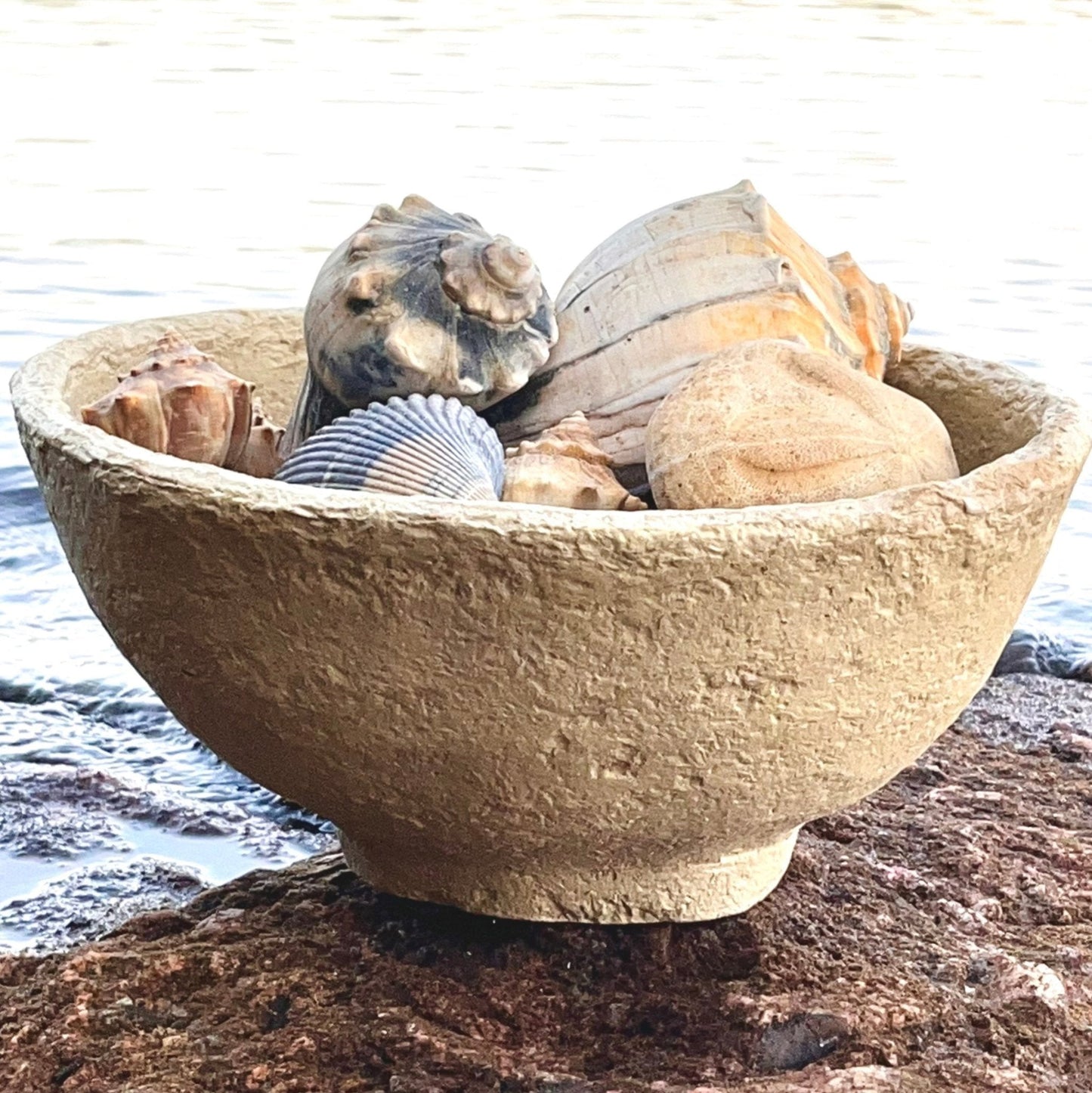 A large oval paper maché bowl with a creamy beige tone, displayed on a rock by a body of water, containing various seashells and pebbles.
